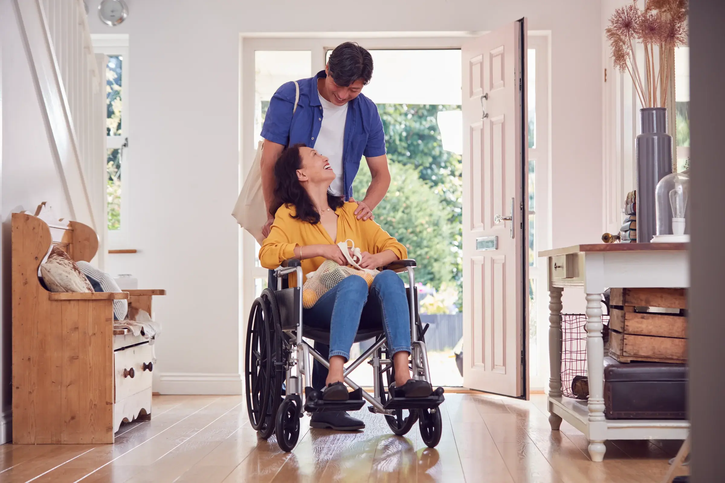 A woman in a wheelchair arriving home through her front door, greeted by her husband